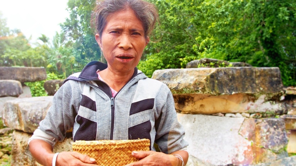  Paní z tradiční vesnice, v ruce kabelku z ratanu, pusu od betele a za sebou megalitické hrobky